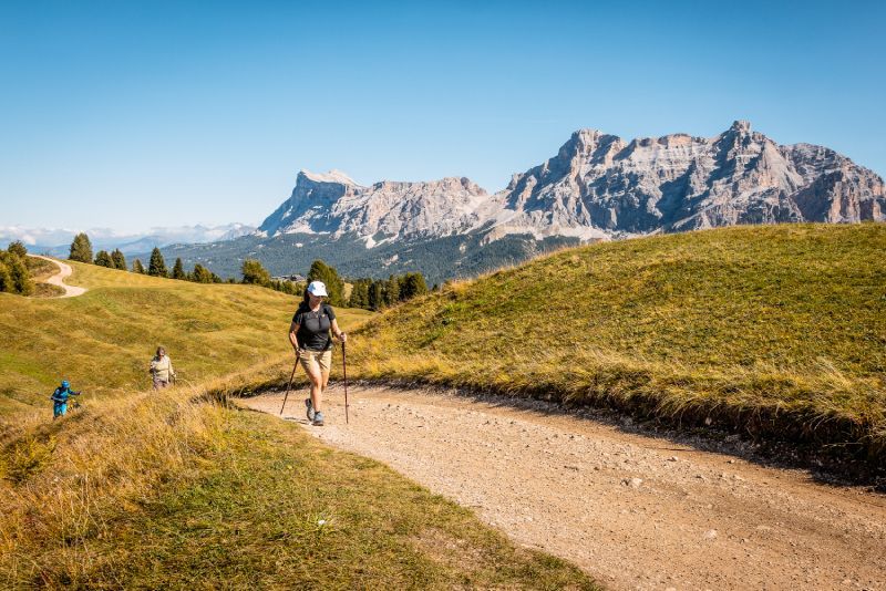 People hiking on a dirt path in a grassy mountain landscape, with mountains in the background under a blue sky.