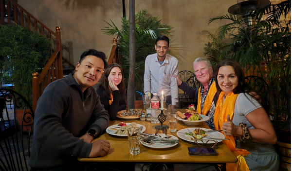 People dining at a restaurant table, with server, food, and plants in the background.