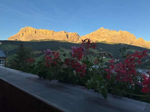 Mountain range lit by golden sunlight, seen from a balcony with blooming red flowers in foreground.