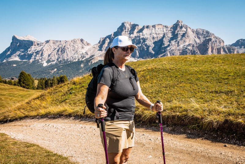 Woman hiking in mountains, using poles, wearing sunglasses and a white hat.