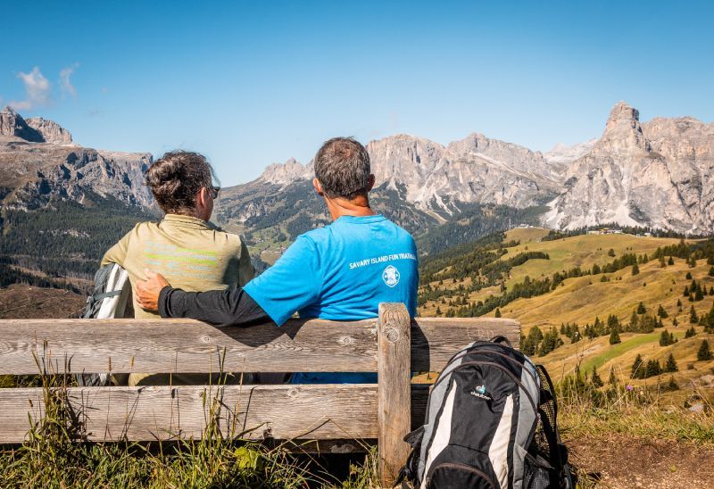 Couple on a bench overlooking mountains; sunny day, backpack in foreground.