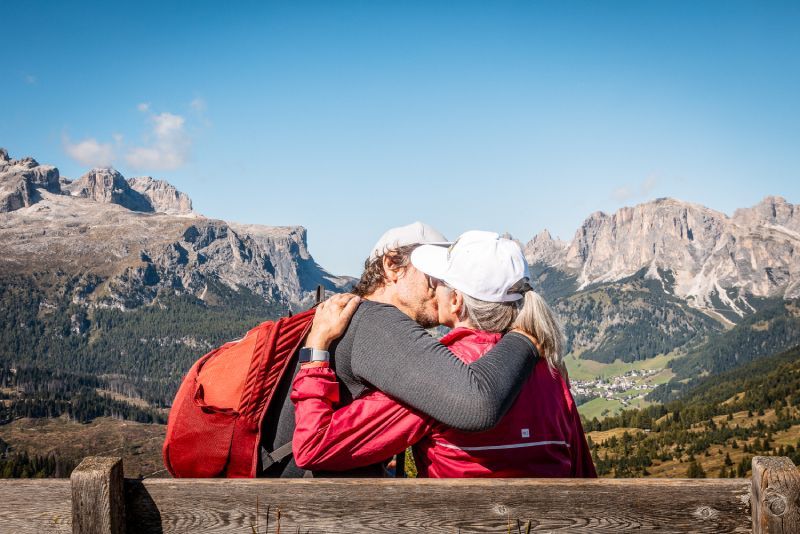 Two men on bikes pose near large, white columns in a park. Sunny day.