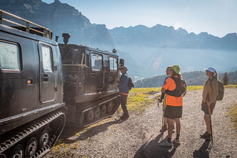 Hikers beside a tracked vehicle on a mountain path, with peaks and valley in the distance.