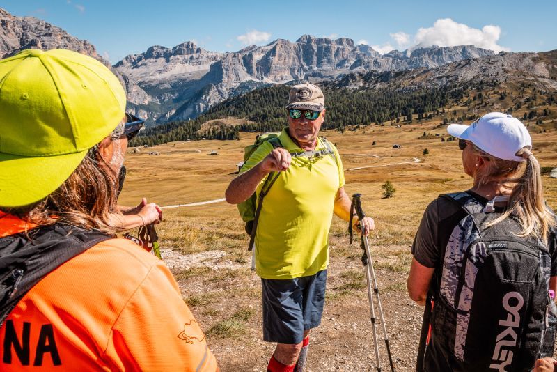 Three people smiling and posing outdoors. Mountains in the background. Man in center with arm around the other two.