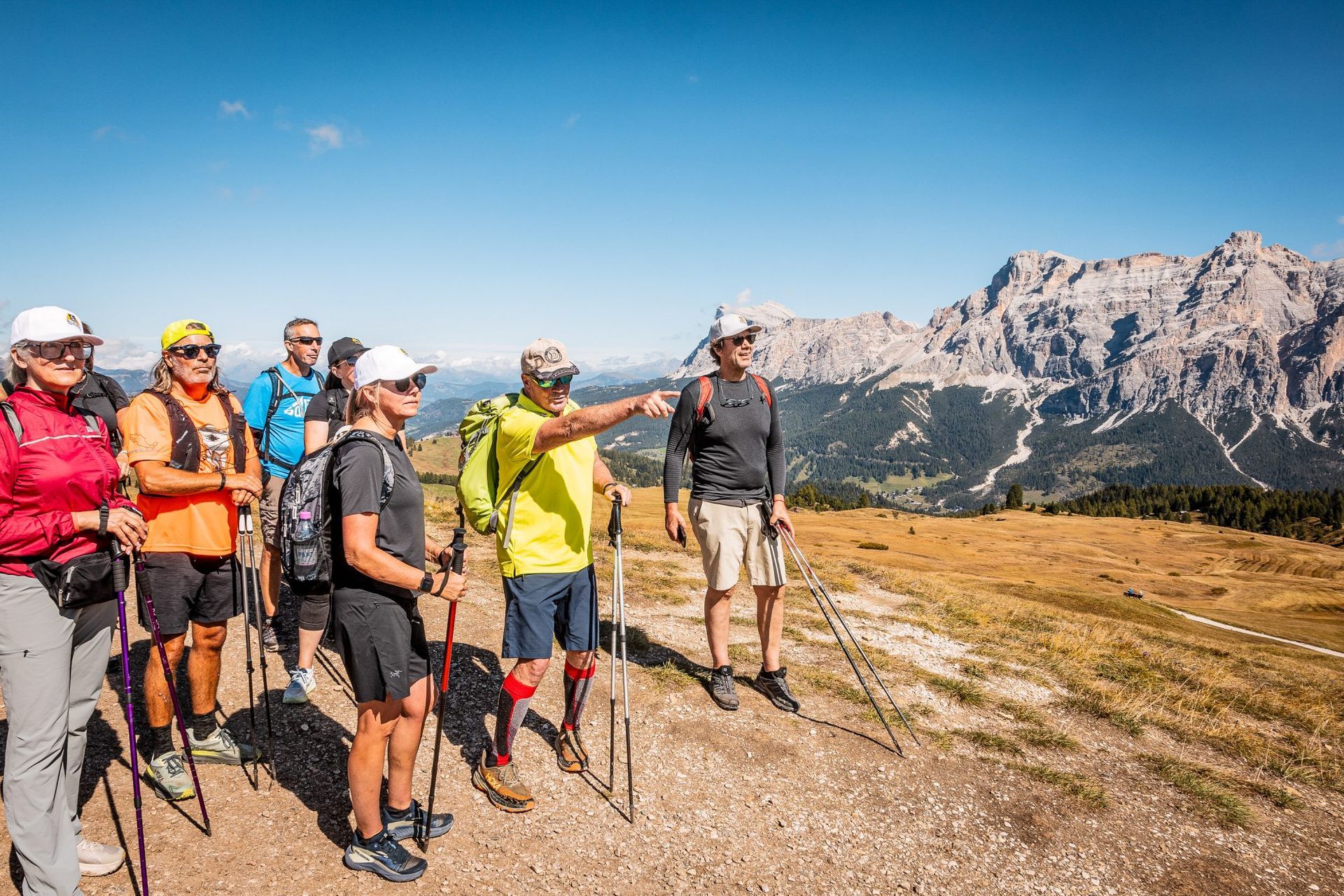 Group hikers on a sunny mountain, pointing towards a scenic vista with a rocky peak.