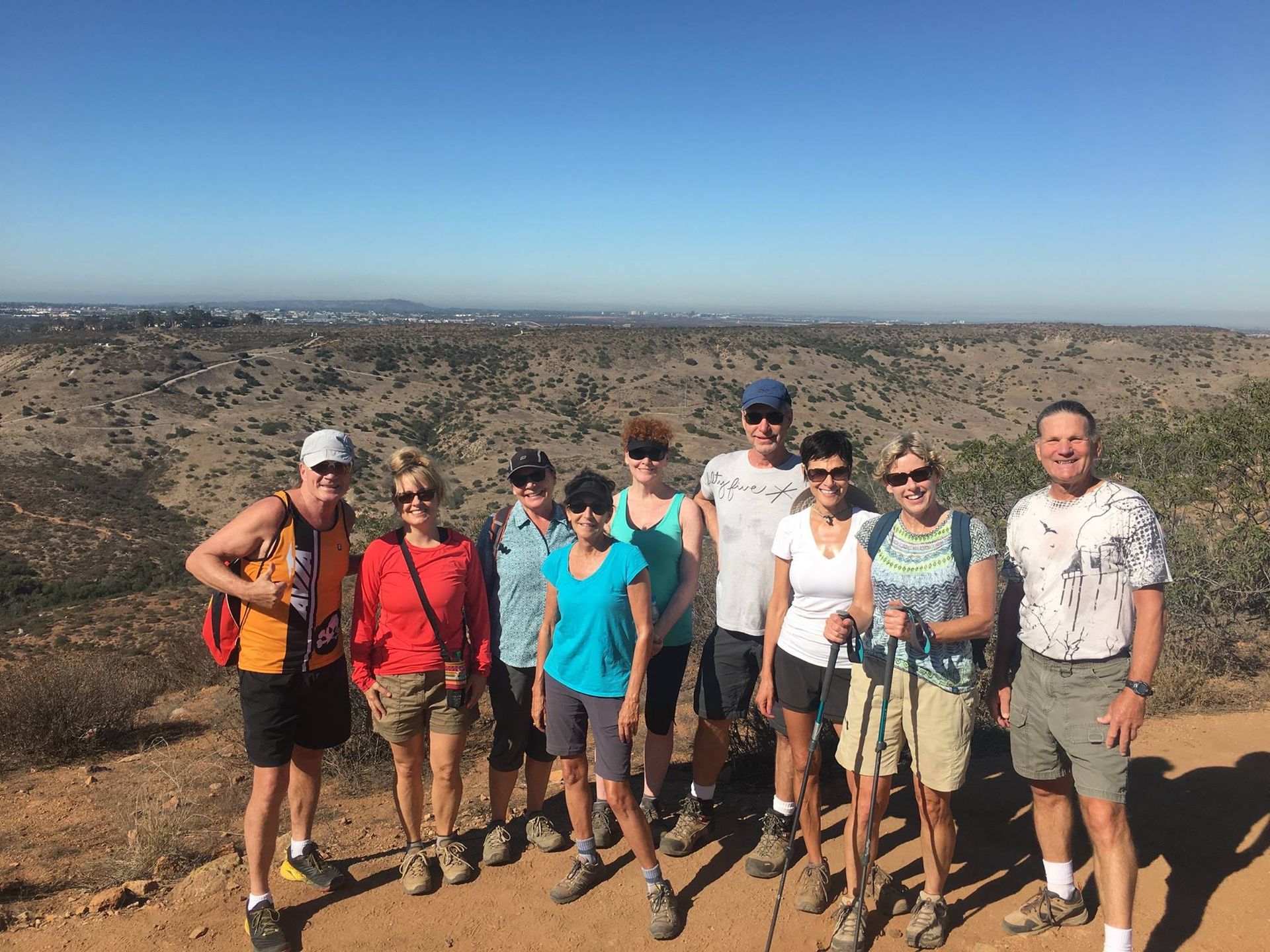 Group of hikers poses on a hilltop overlooking a dry, hilly landscape under a blue sky.