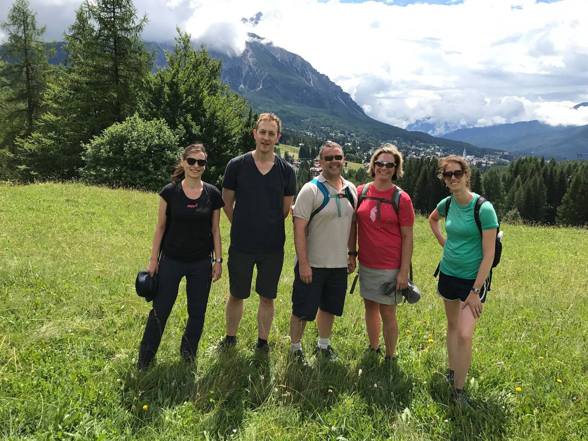 Five people standing in a grassy field with mountains in the background. Sunny day.