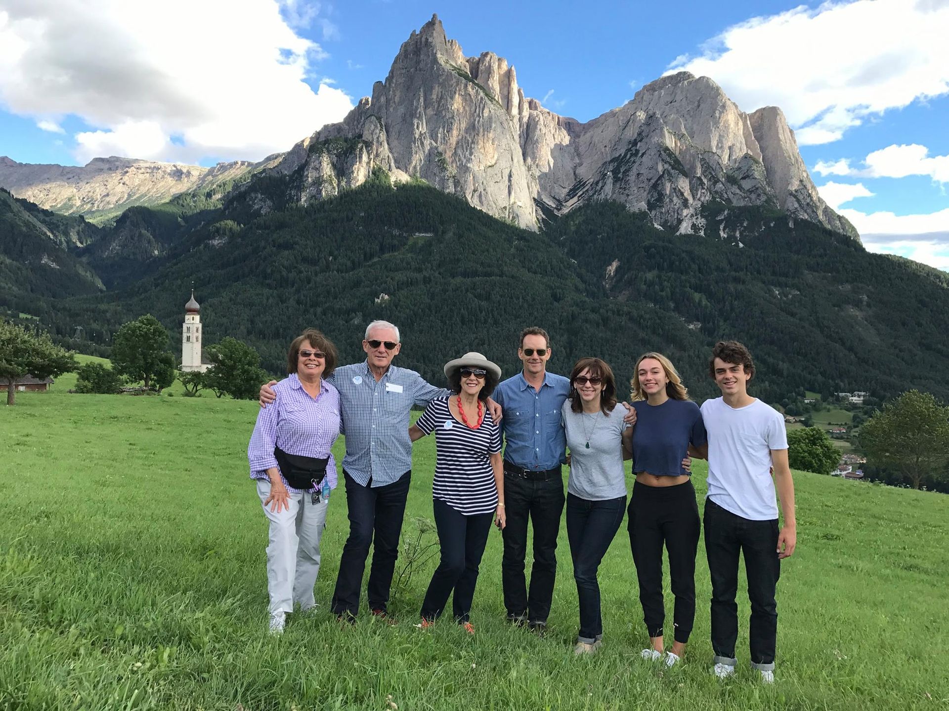 Group of people smiling in front of a mountain range. Green grass, blue sky.