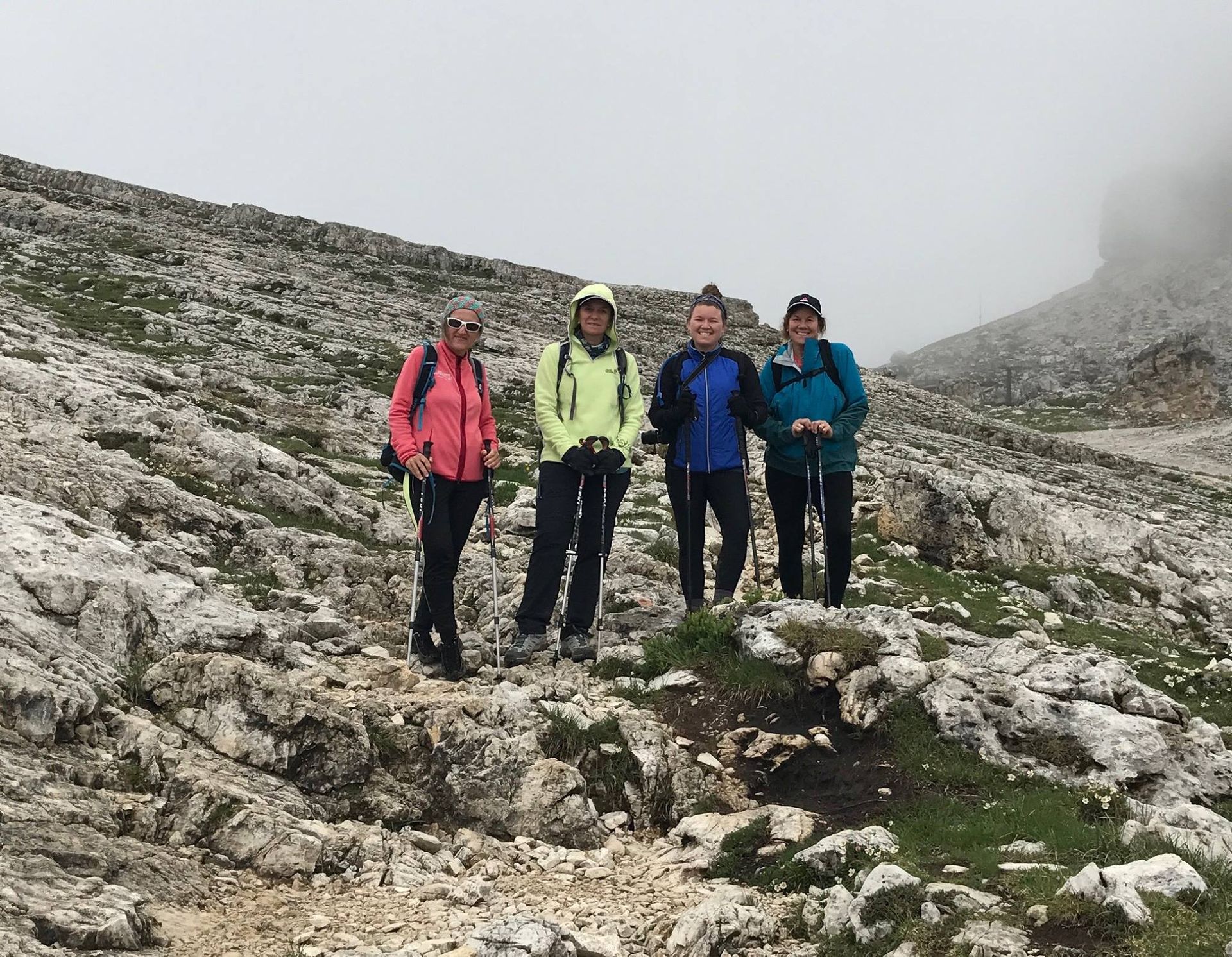 Four women hikers stand on a rocky mountain path. They wear colorful jackets and use hiking poles. Overcast sky.
