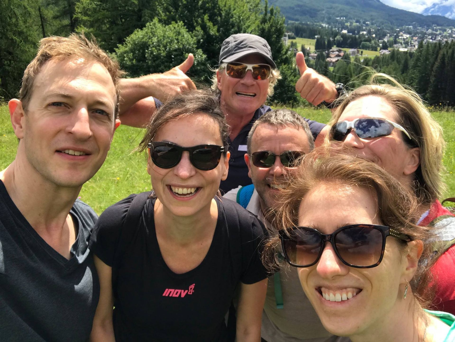 Group of smiling people in sunglasses, thumbs up, outdoors in a grassy field with a town visible.
