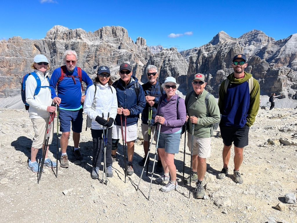Group of hikers pose on a mountain peak with rocky peaks in the background under a clear blue sky.