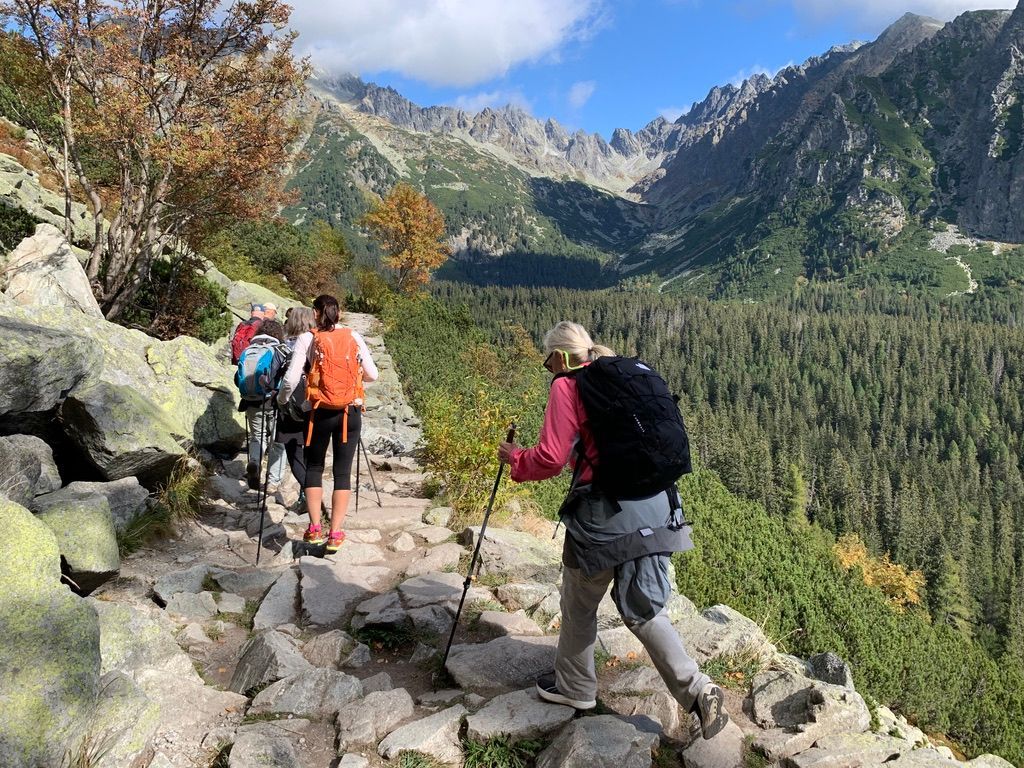 Hikers with backpacks on a rocky trail in a mountain landscape, under a blue sky.