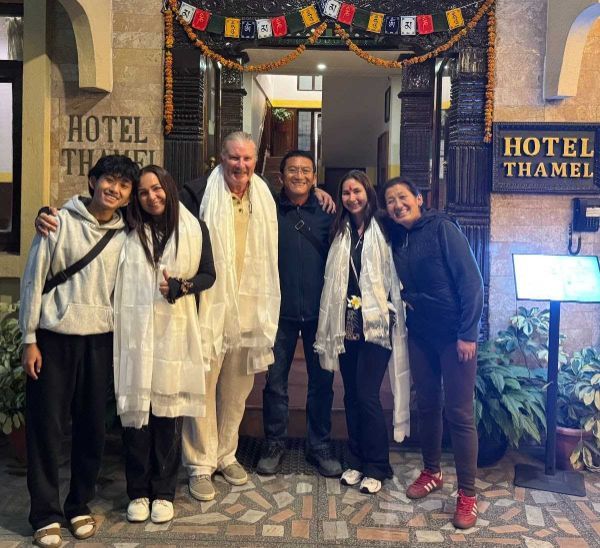 Group of six people posing outside Hotel Thamel entrance. Everyone smiles, some draped in white scarves.