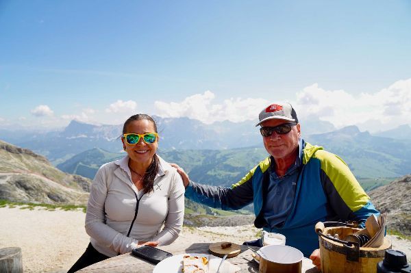 Couple at outdoor table with mountain view; sunny day. Woman in sunglasses smiles, man's arm around her.