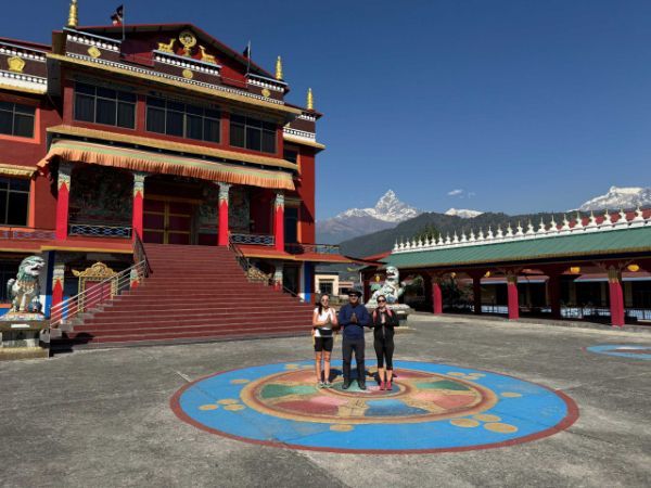 Three people standing in front of a red Buddhist temple with mountains in the background. Sunny day.