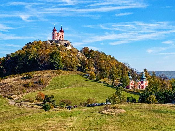 Red church with two towers atop a hill, surrounded by trees and a green meadow, under a blue sky.