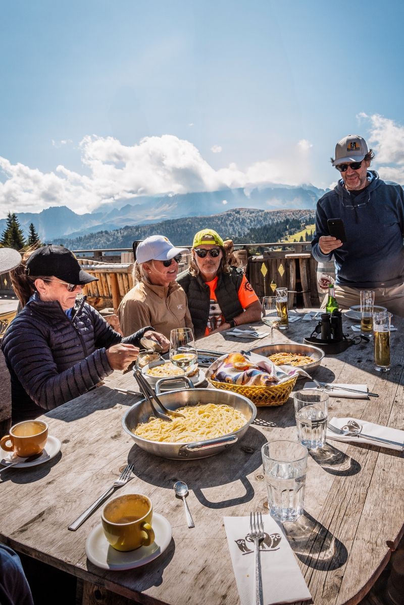 People enjoying a meal at a wooden table on a mountainside; sun shining; food visible; blue sky.