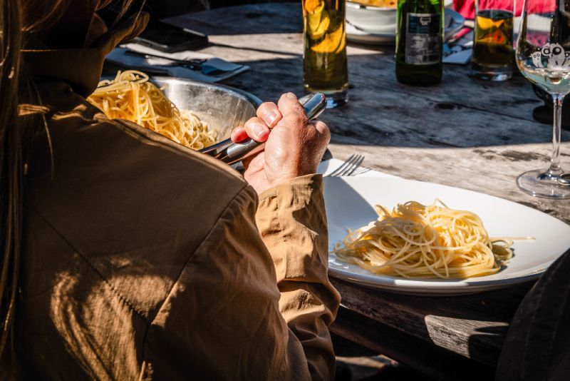 Person eating spaghetti outdoors at a wooden table with drinks and other dishes.