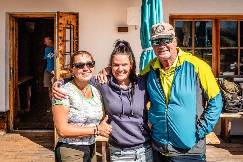 Group of seven people posing in front of a monument and mountains; green grass, trees, blue sky.