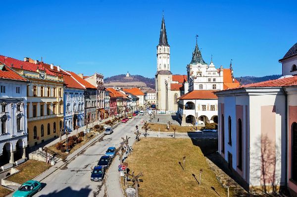 Street view of a European town with colorful buildings, cars, and a church tower under a blue sky.