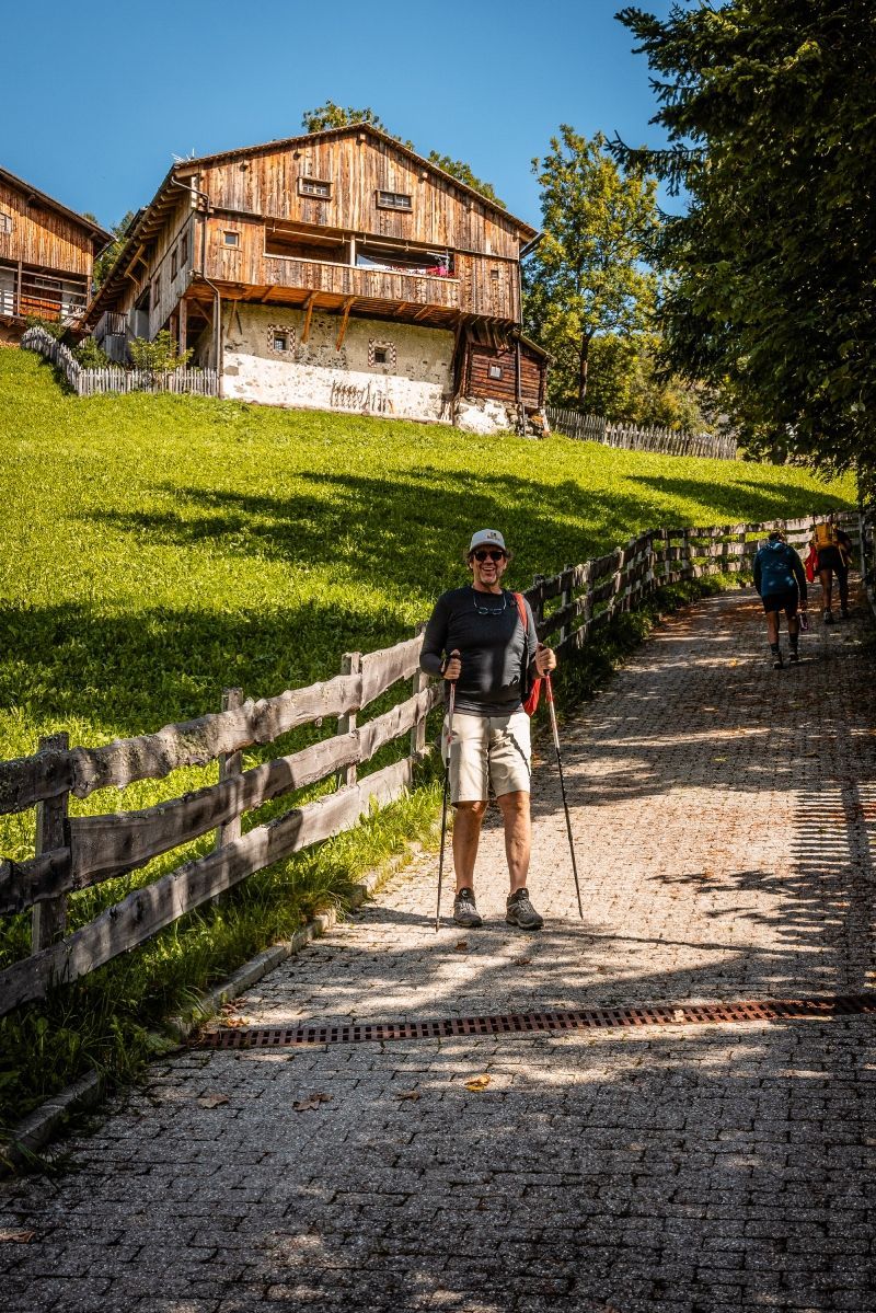 Man hiking on a path with wooden buildings on a grassy hill. Sunny day.