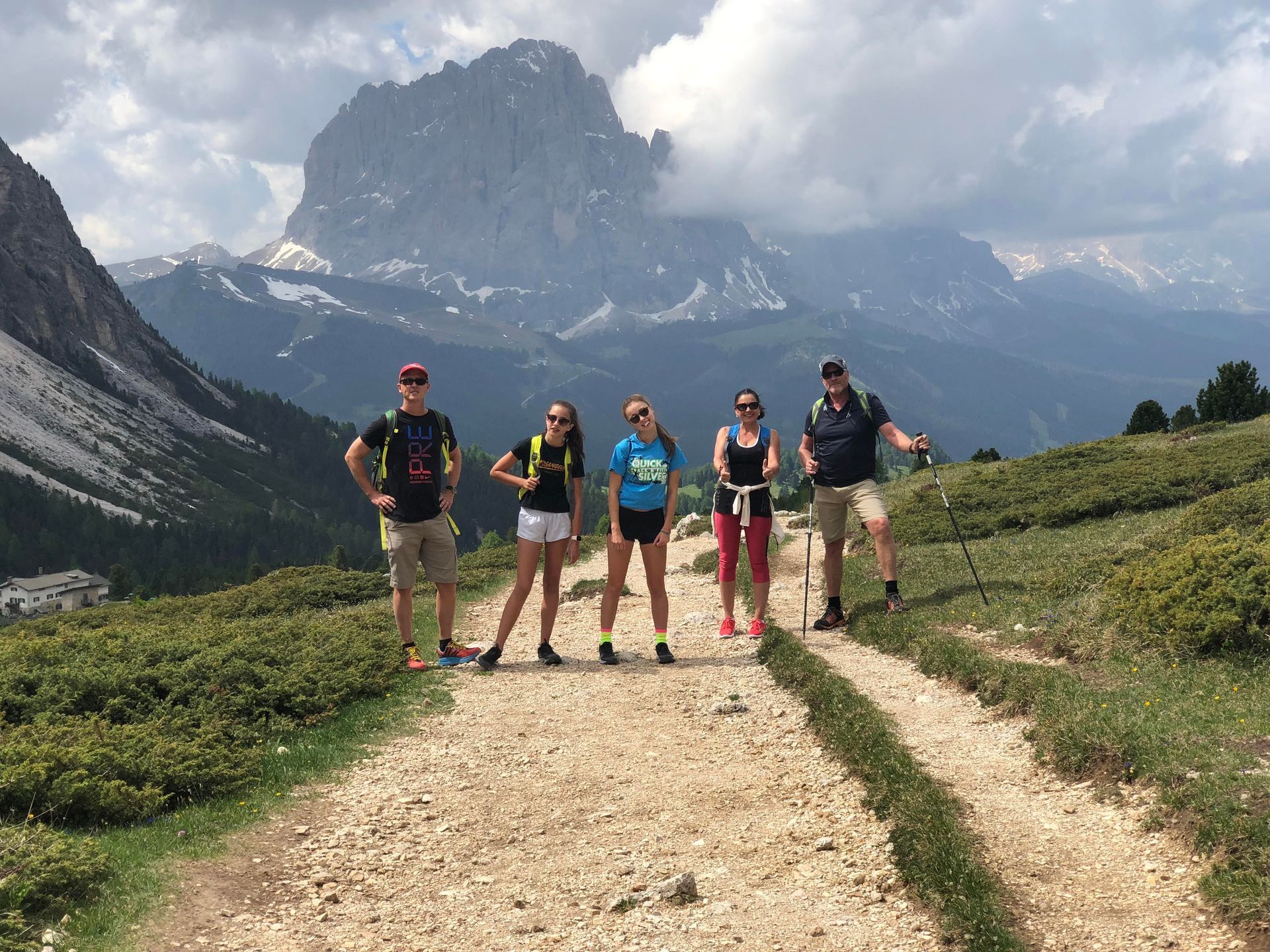 Five hikers on a dirt path in front of mountains, cloudy sky.