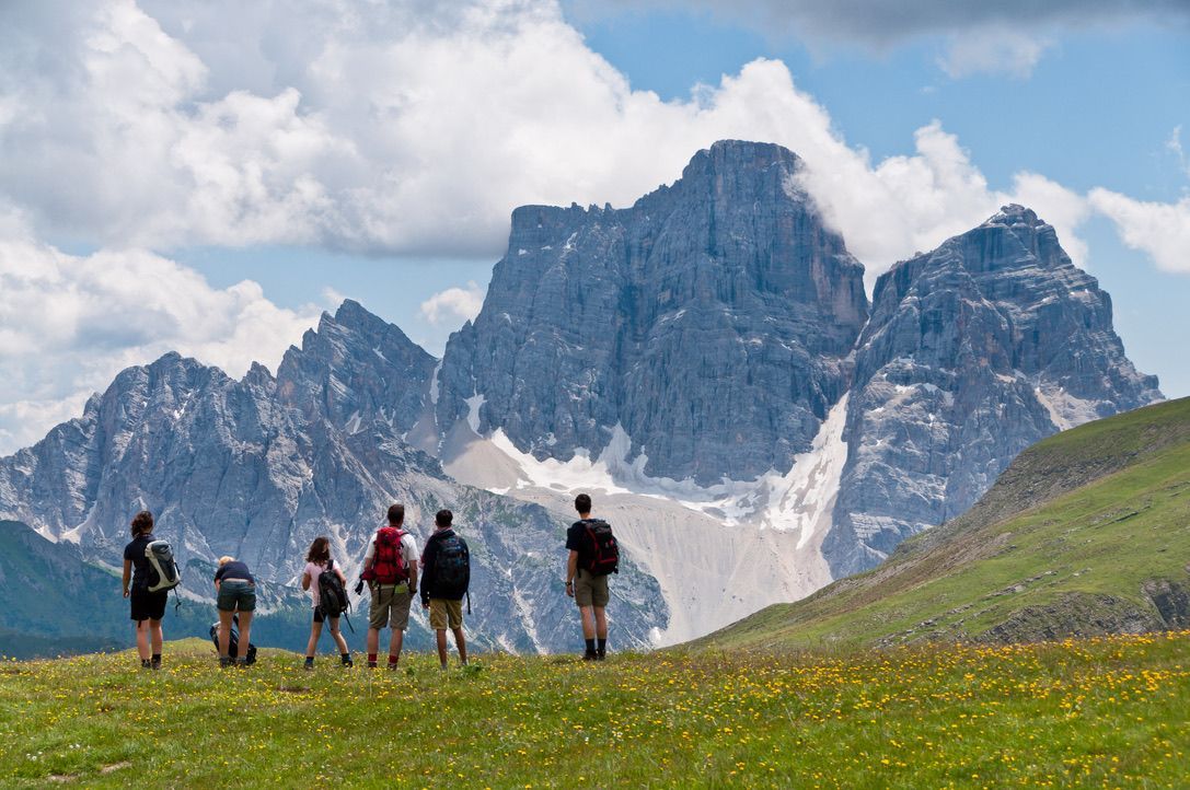 Hikers in a grassy field, gazing at a large, rocky mountain under a cloudy sky.