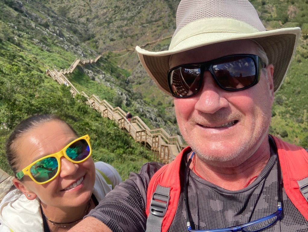 Couple smiling on a mountain trail with wooden stairs; man in hat, woman in yellow sunglasses.