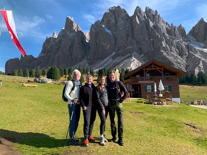 Four people pose in front of mountains and a cabin on a grassy slope.