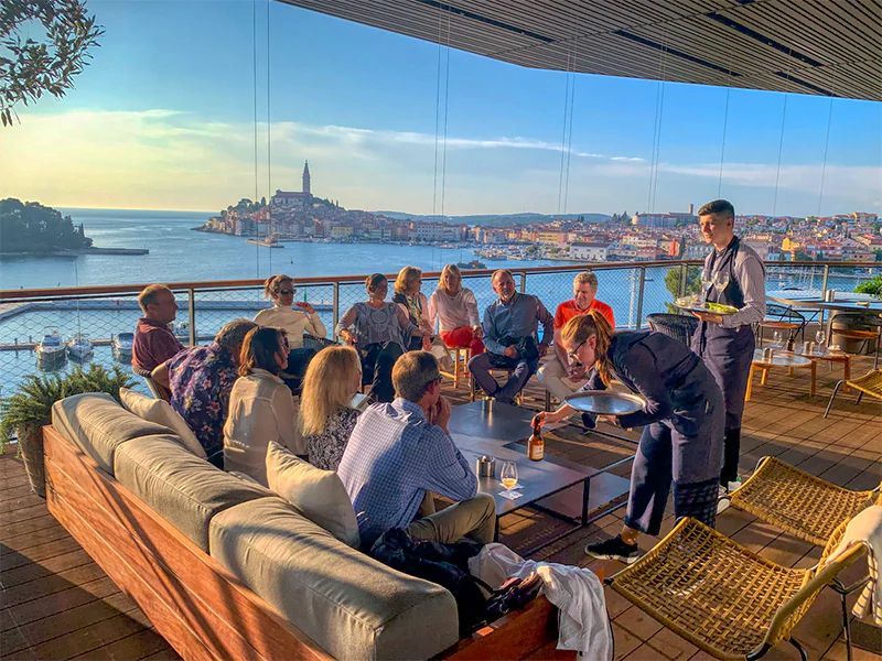 People on a rooftop patio overlooking a coastal city, served drinks by staff; sunny day.