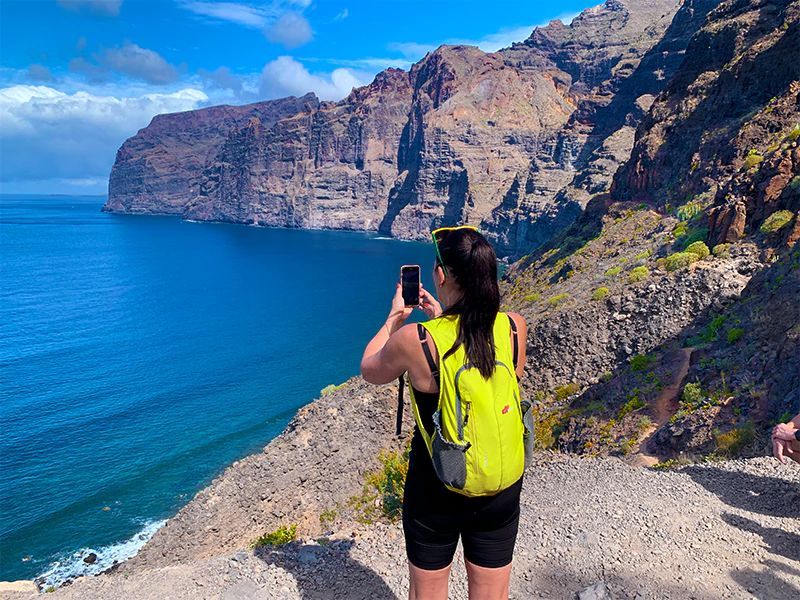 Woman taking photo of cliffs over ocean, wearing yellow backpack, black shorts, and top.