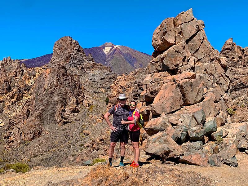 Man and woman stand on a rocky mountain trail, with the peak of a volcano in the background under a clear blue sky.