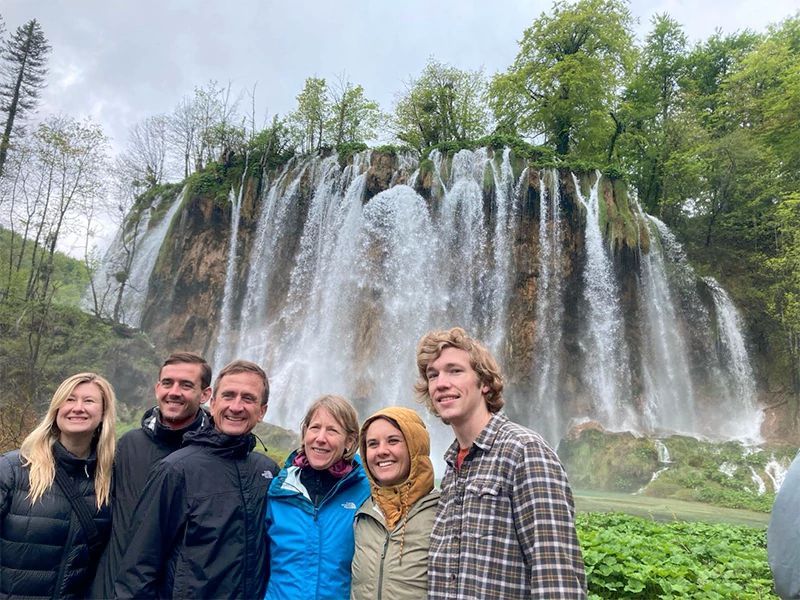 Group of six people standing in front of a large waterfall, trees behind them. Cloudy day.