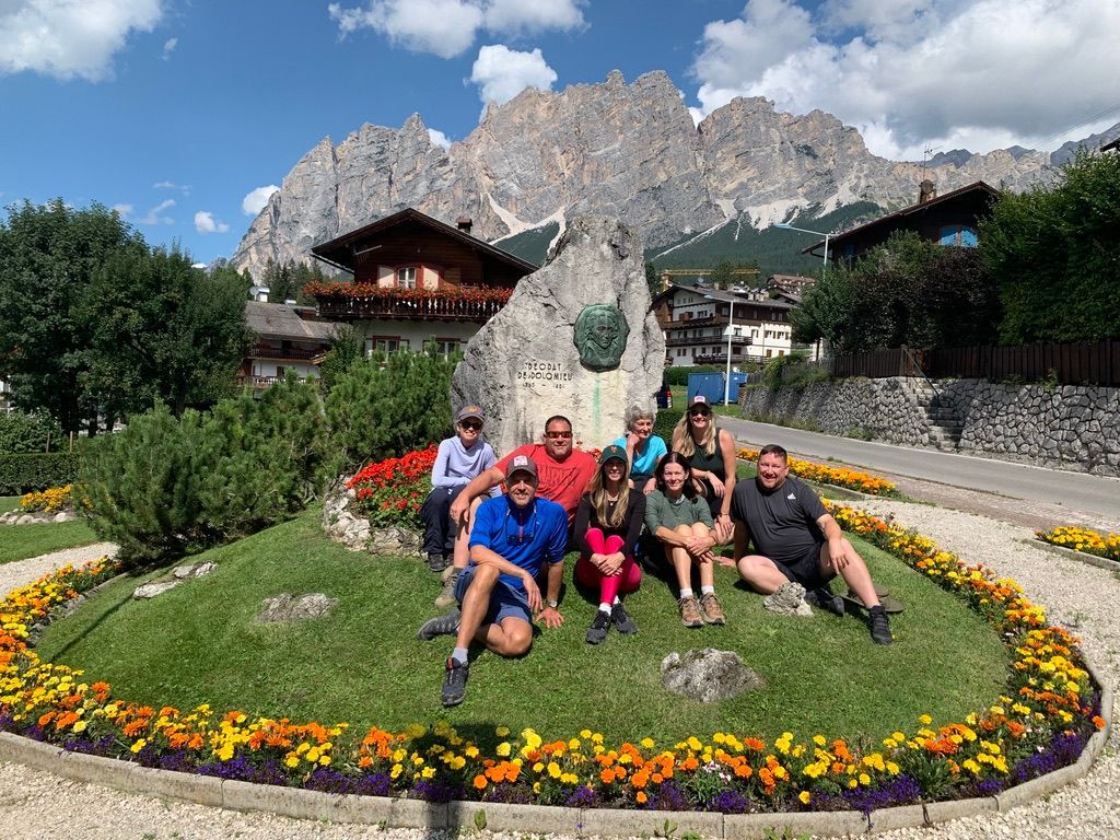 Group of people pose by a stone monument with mountains in the background, flowers surround the grassy area.
