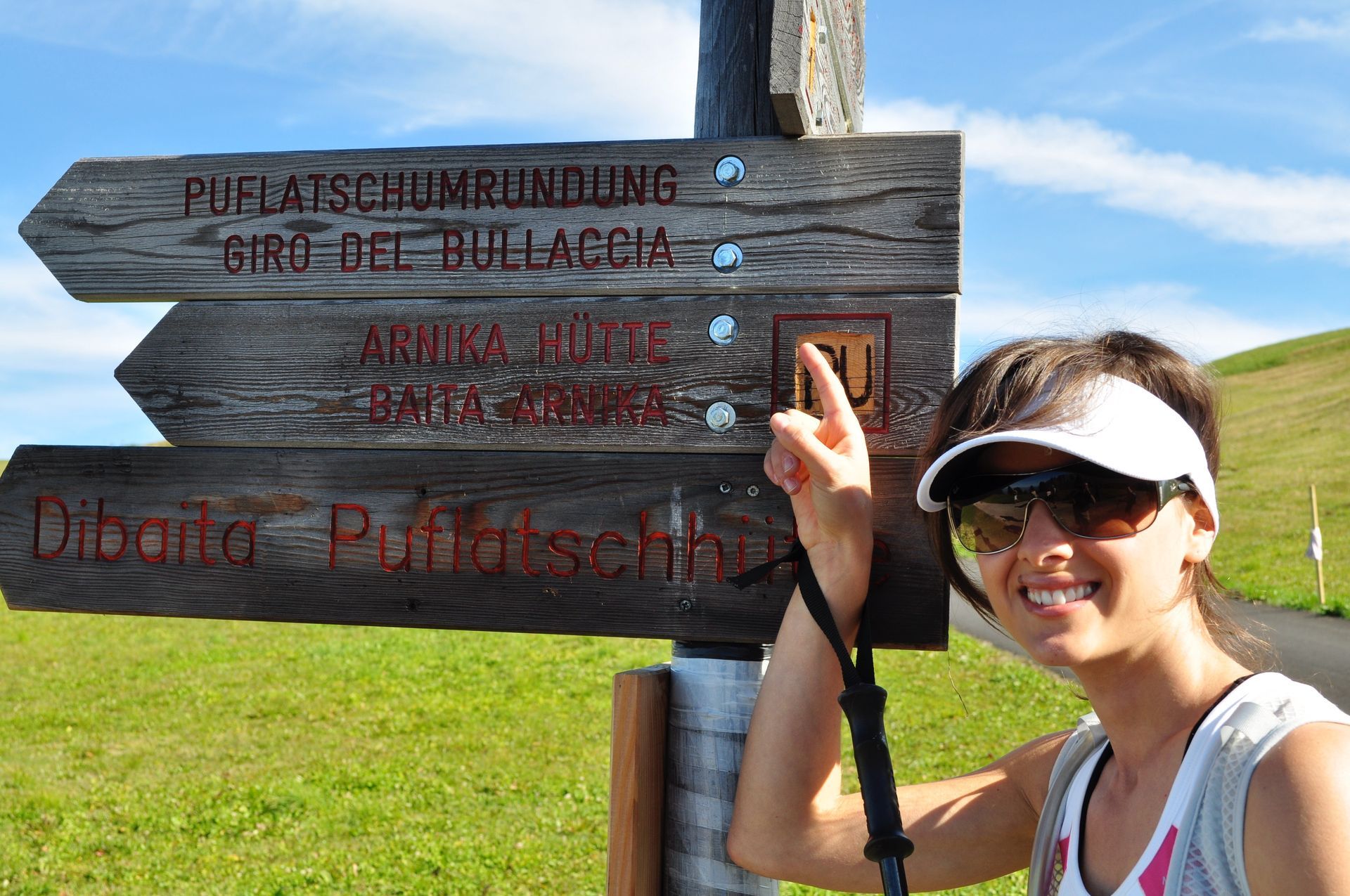 Woman points to a wooden trail sign in a grassy, mountainous setting.