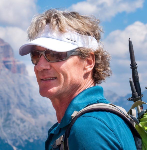 Man hiking in mountains, wearing sunglasses and visor, with backpack.
