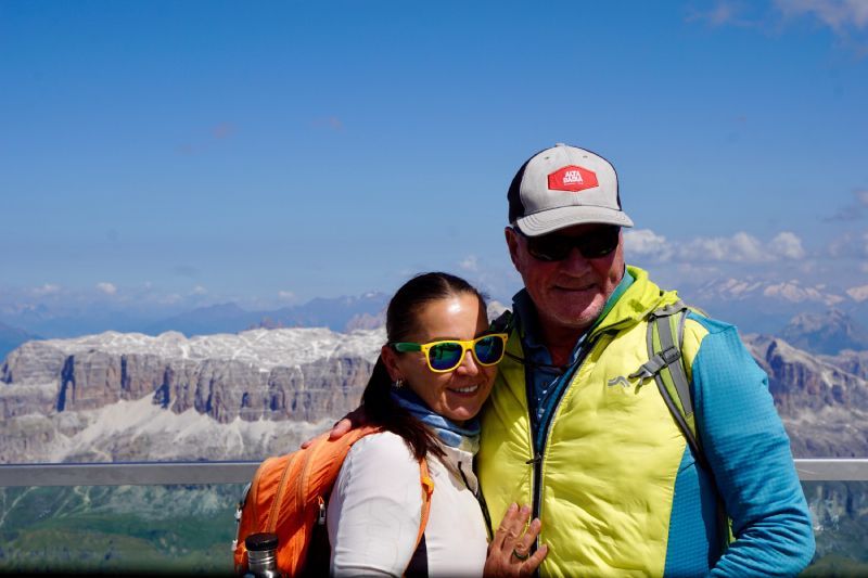 A couple smiles at the camera, posing against a mountain range. They wear sunglasses, a cap, and a puffy vest.