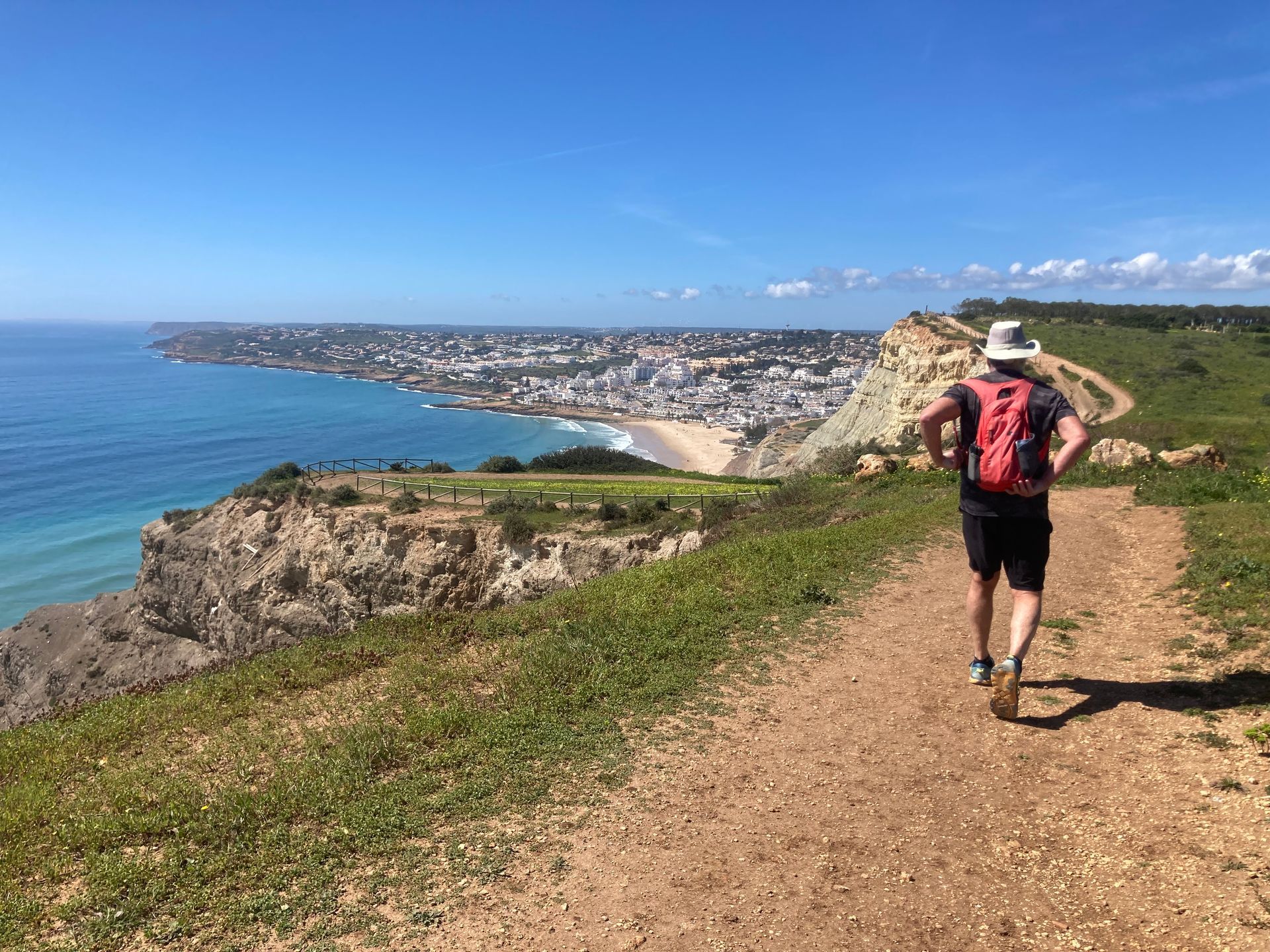 Man hikes on a coastal trail with a town and ocean view under a bright blue sky.