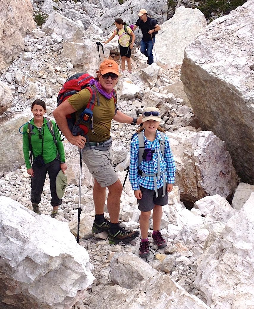 Group hiking rocky terrain; sunny day. Woman in orange hat, backpack, and two children pose for photo.