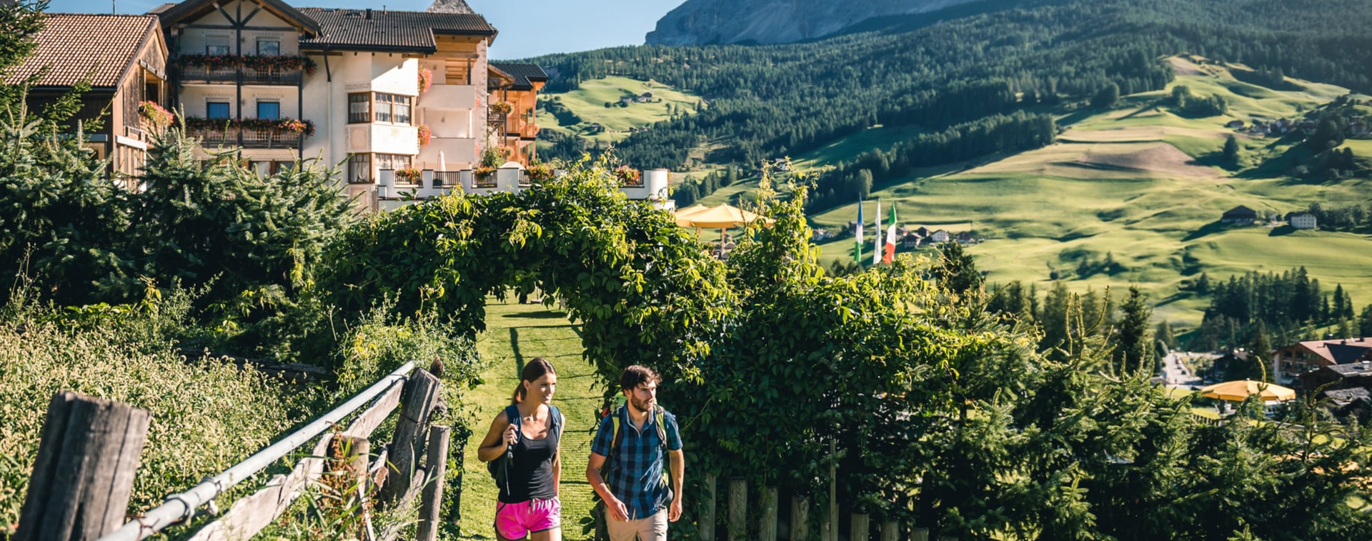 Couple walks along a path lined with greenery, a village and mountains in the background.