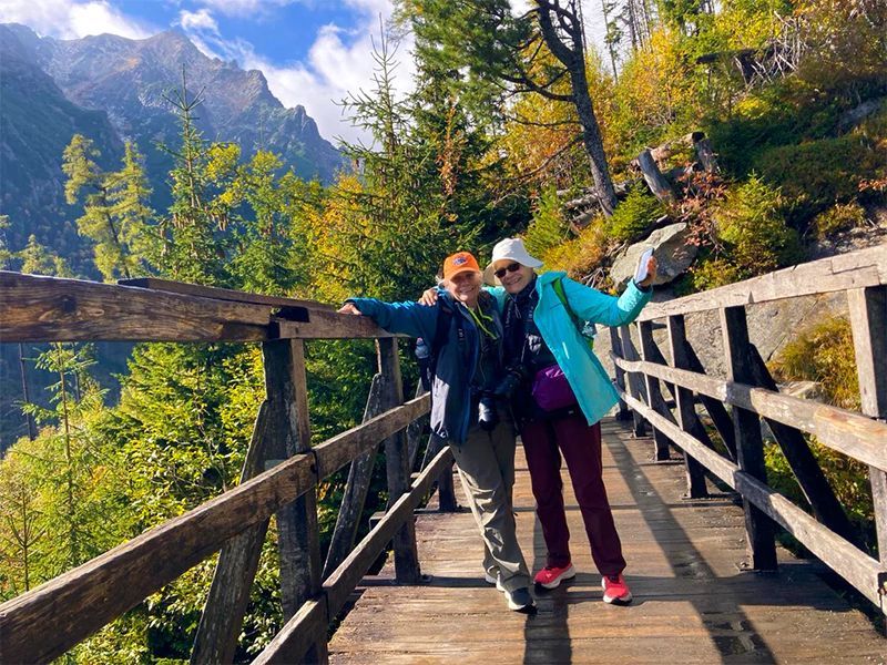 Two people on a wooden bridge, arms out, smiling. Mountains and fall foliage in background.