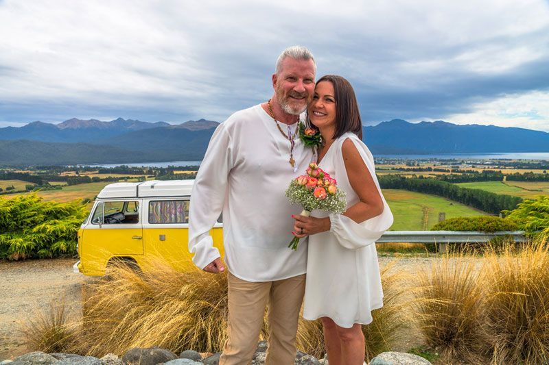 Couple, smiling, pose for a wedding photo. They stand before a yellow VW van, scenic view of mountains and fields.