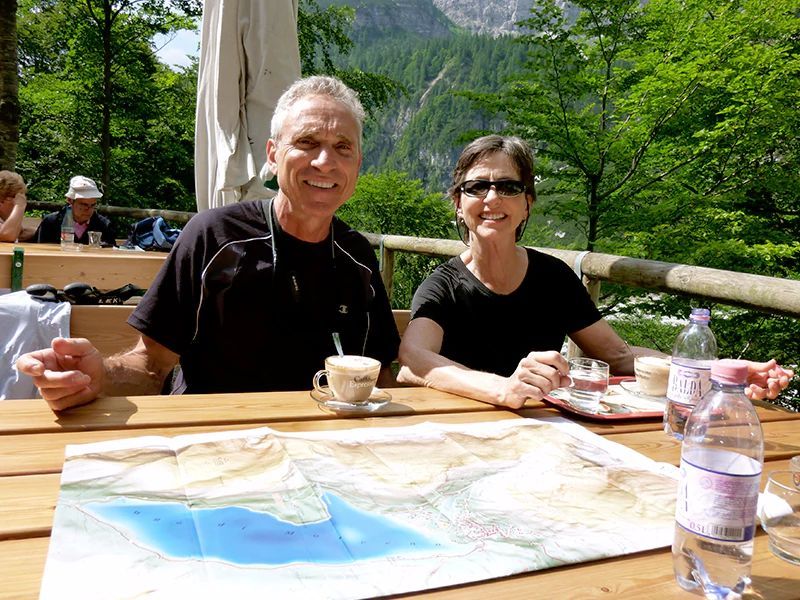 Couple at a table with a map, drinks, and mountain view. Smiling and posing outdoors.