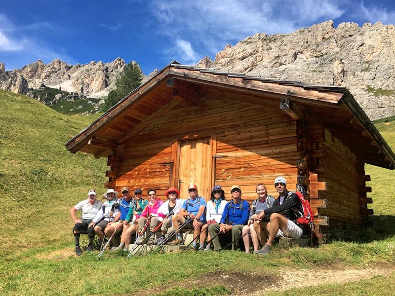 Group of people sits in front of a wooden cabin in the mountains. Sunny day.