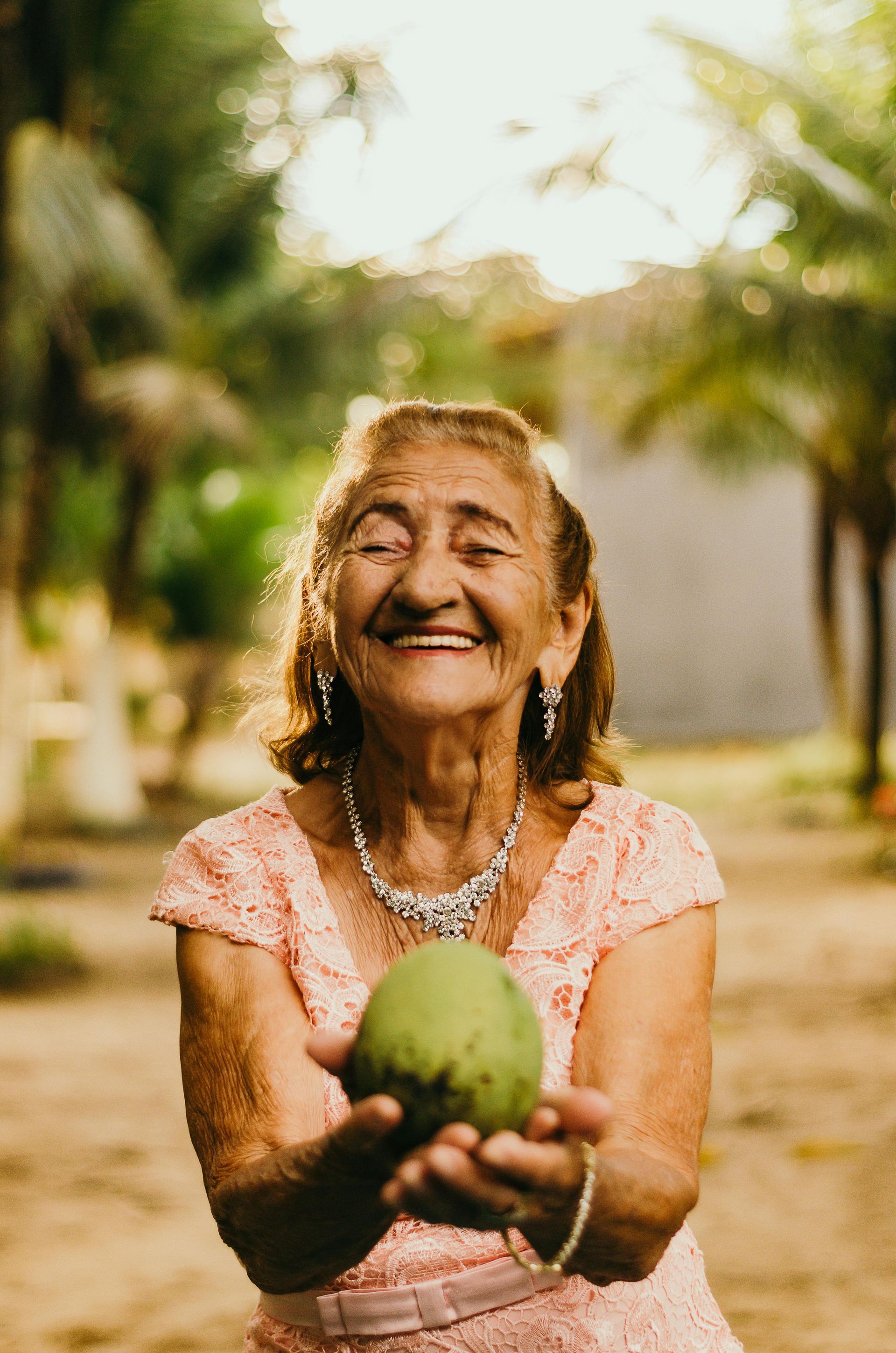 Persona sonriente sosteniendo una fruta verde al aire libre, vistiendo un vestido de encaje rosa y joyas, fondo soleado.
