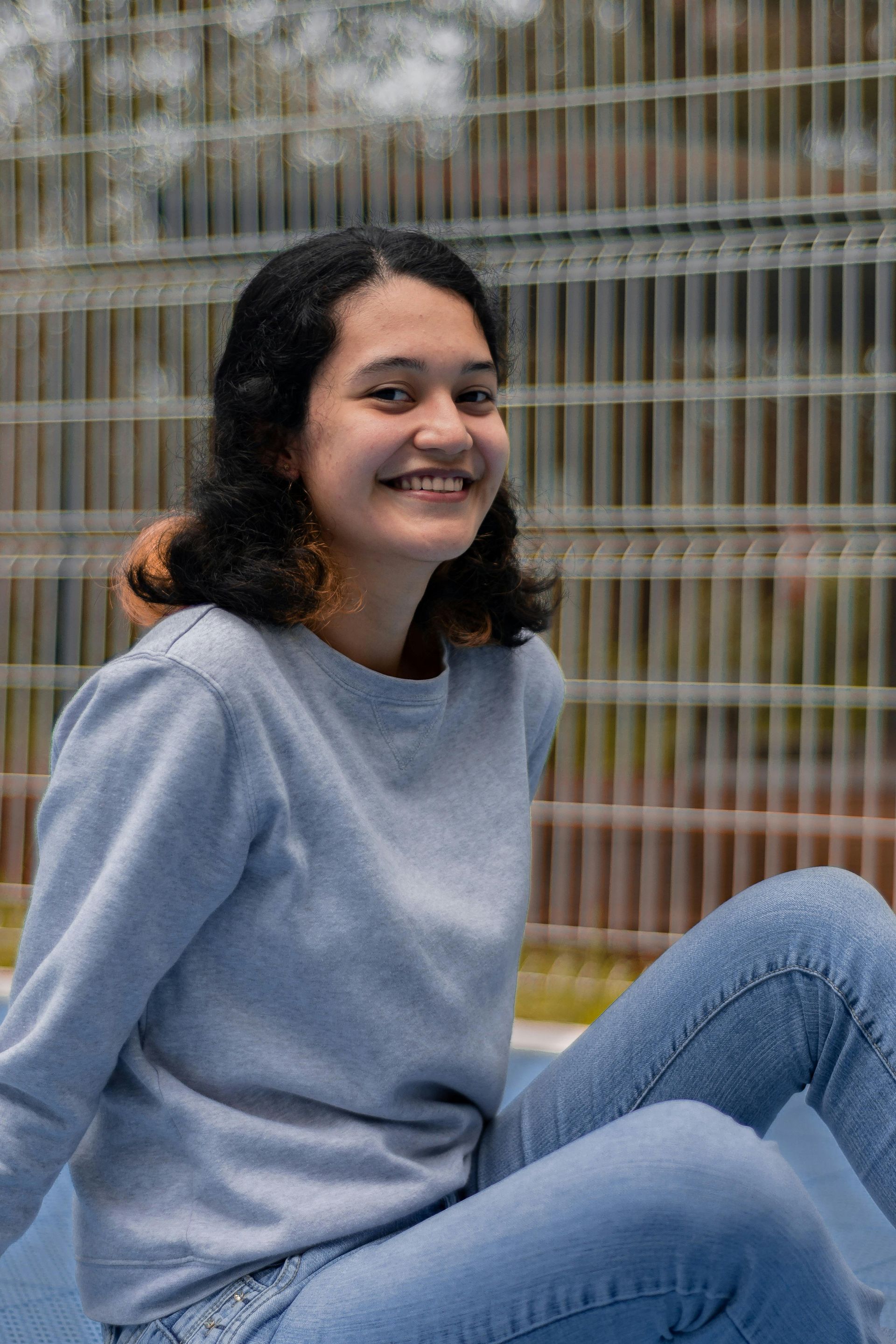 Una mujer con sudadera azul y vaqueros sonríe mientras está sentada al aire libre cerca de una valla.