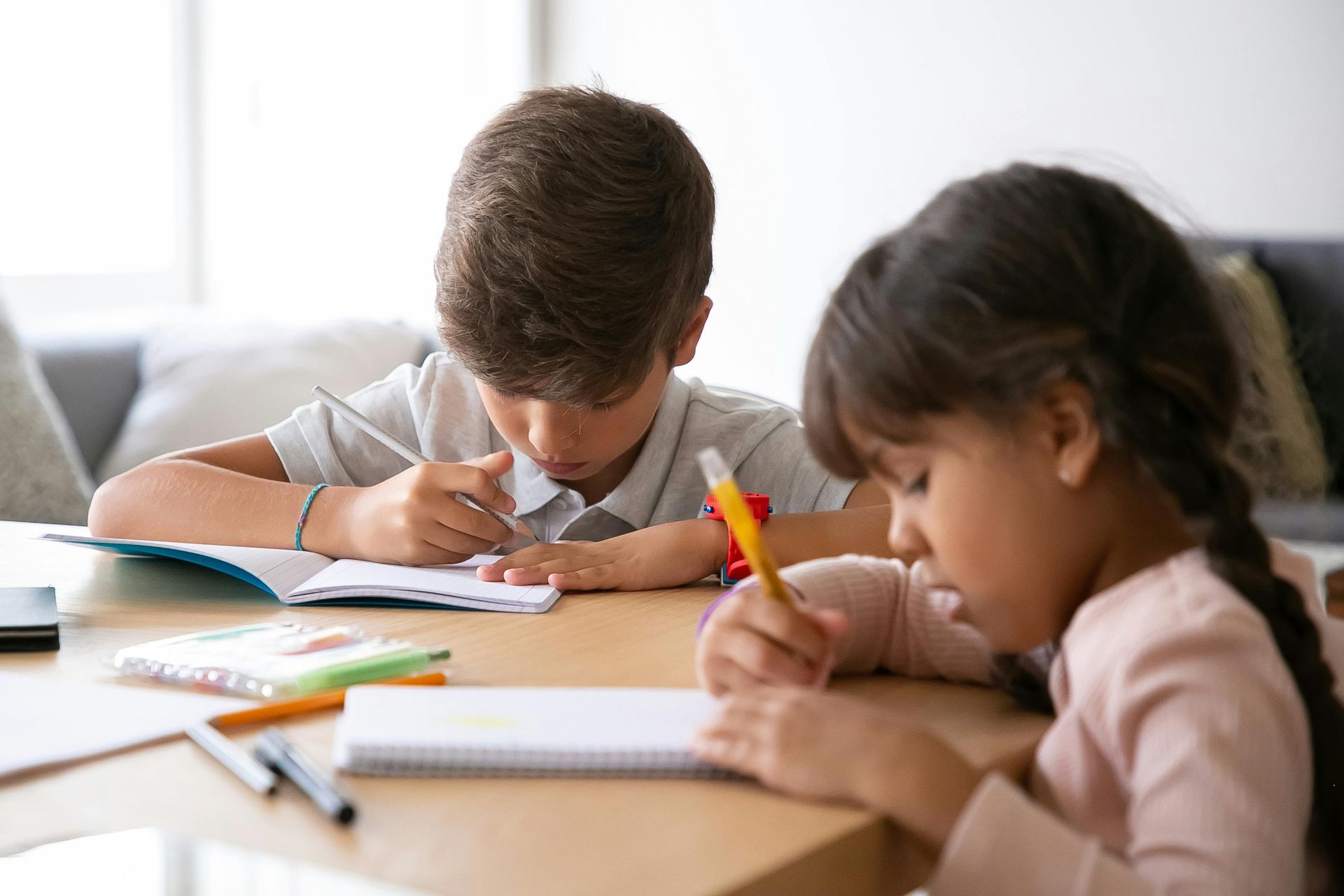 Dos niños, concentrados en escribir en cuadernos sobre una mesa de madera. Se ven lápices y bolígrafos.