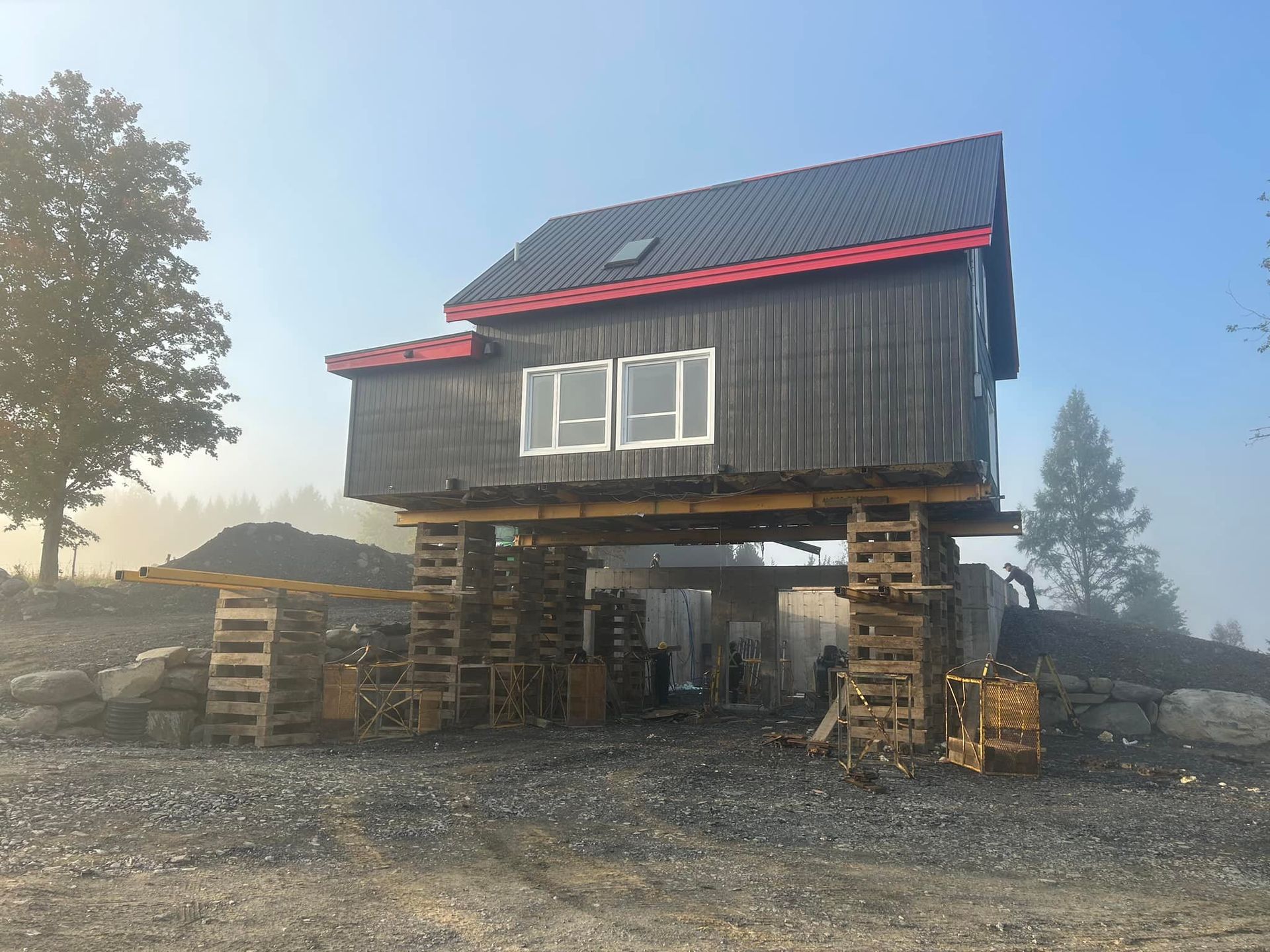Une maison en bois sombre est surélevée sur des supports en bois, un chantier de construction sur un terrain gravillonné.