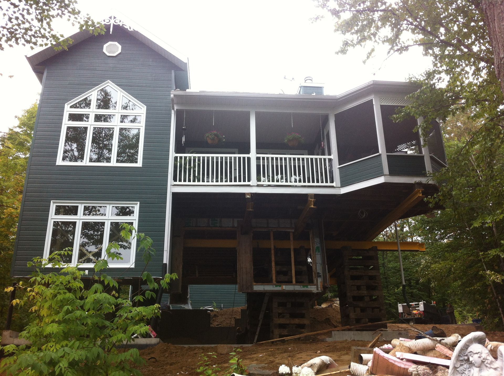 Maison verte aux finitions blanches, porche surélevé et grandes fenêtres, nichée au milieu des arbres.