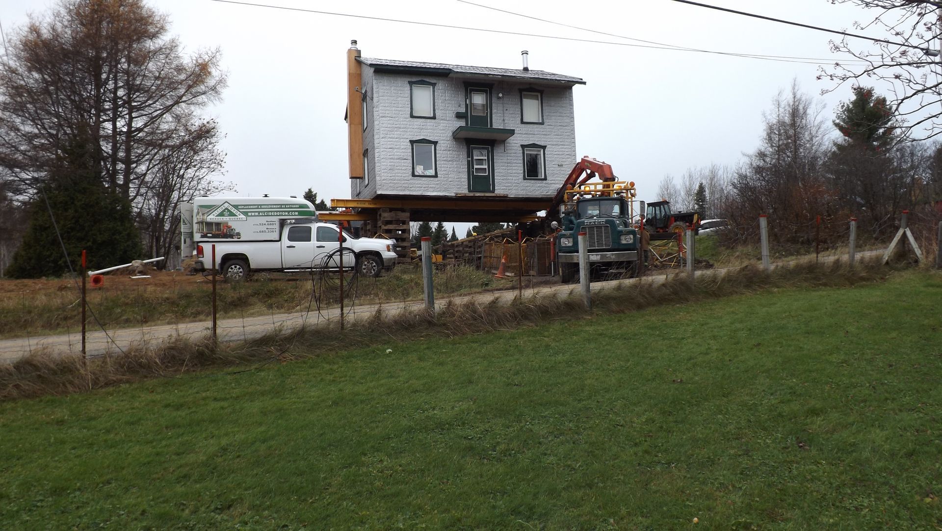 La maison est déplacée, soulevée par des étais. Un camion blanc et des engins de chantier sont présents sur la pelouse.